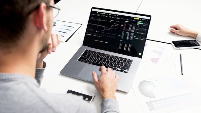 Alt-text: Visão por cima do ombro de um profissional de marketing analisando dashboards com gráficos de desempenho financeiro em um laptop. A mesa branca de escritório está organizada com papéis de relatórios, um tablet e um smartphone, em um ambiente moderno e bem iluminado. 